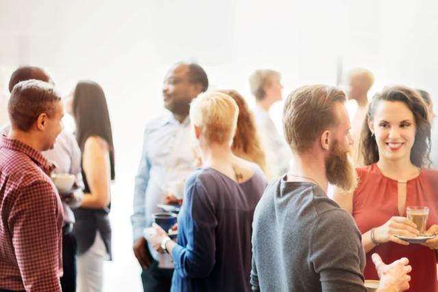 diverse group of people stand around talking, some with coffee in their hands